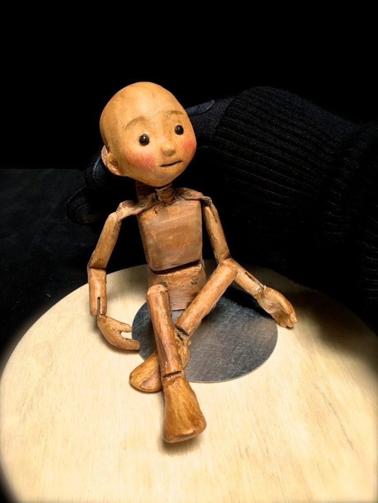La petite marionnette ME, toute menue, articulée et faite en bois, est assise sur son présentoir (plaque de métal collée sur une platefoirme de bois).